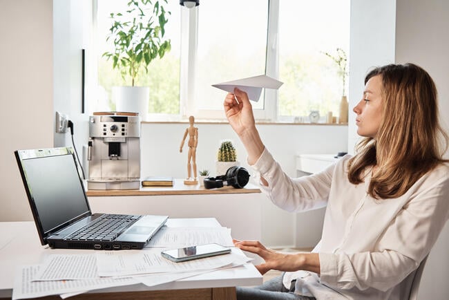 Woman sitting at a desk holding a paper airplane while working on a laptop, with papers spread out in front of her.