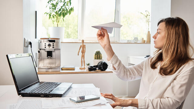 Woman sitting at a desk holding a paper airplane while working on a laptop, with papers spread out in front of her.